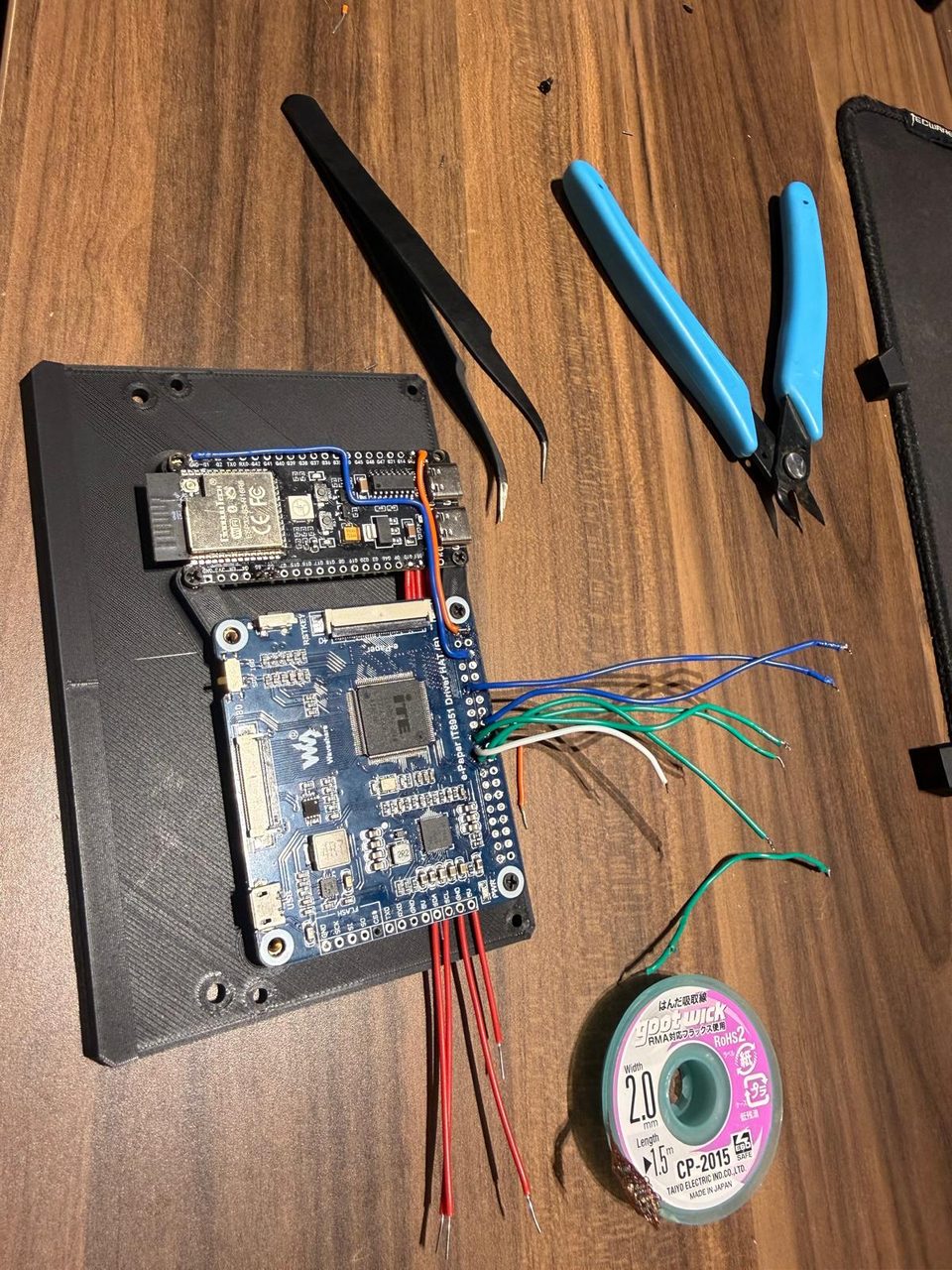 The ESP32-S3 board and the IT8951 driver board mounted on a black plate, surrounded by flush cutters, tweezers and solder wick, with coloured wires half-attached.